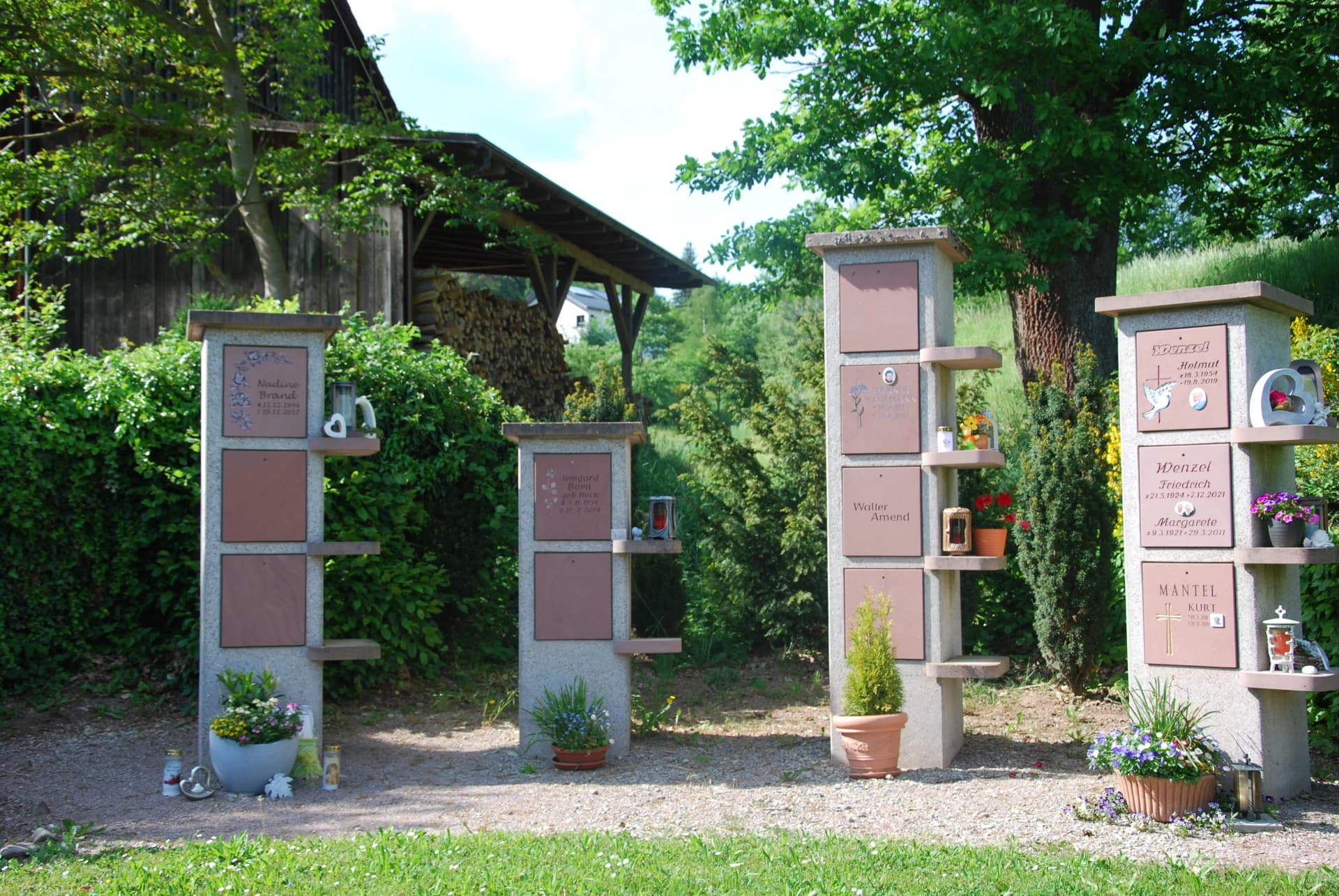 Steinstelen mit Namenstafeln auf einem Friedhof, umgeben von Blumenschmuck und Grabkerzen