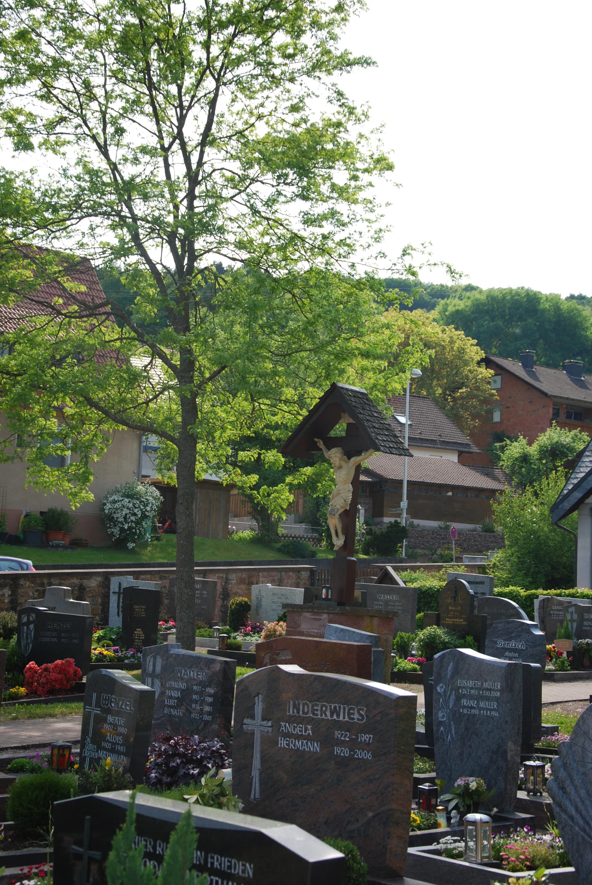 Friedhof mit Grabsteinen, Blumenschmuck, Bäumen und einem überdachten Kreuz mit Kruzifix