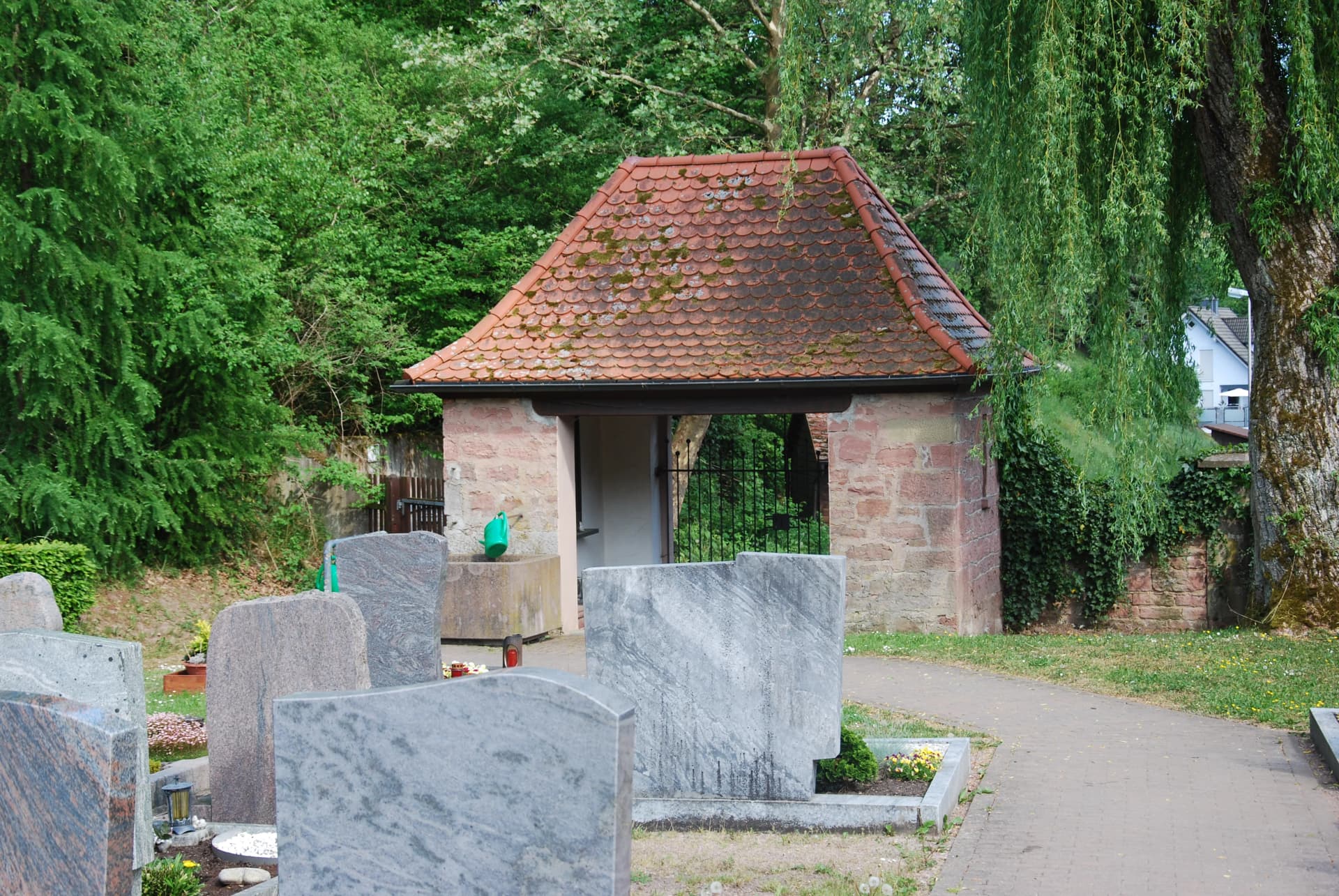 Kleines Torhaus mit Ziegeldach auf einem Friedhof, umgeben von Bäumen und Grabsteinen