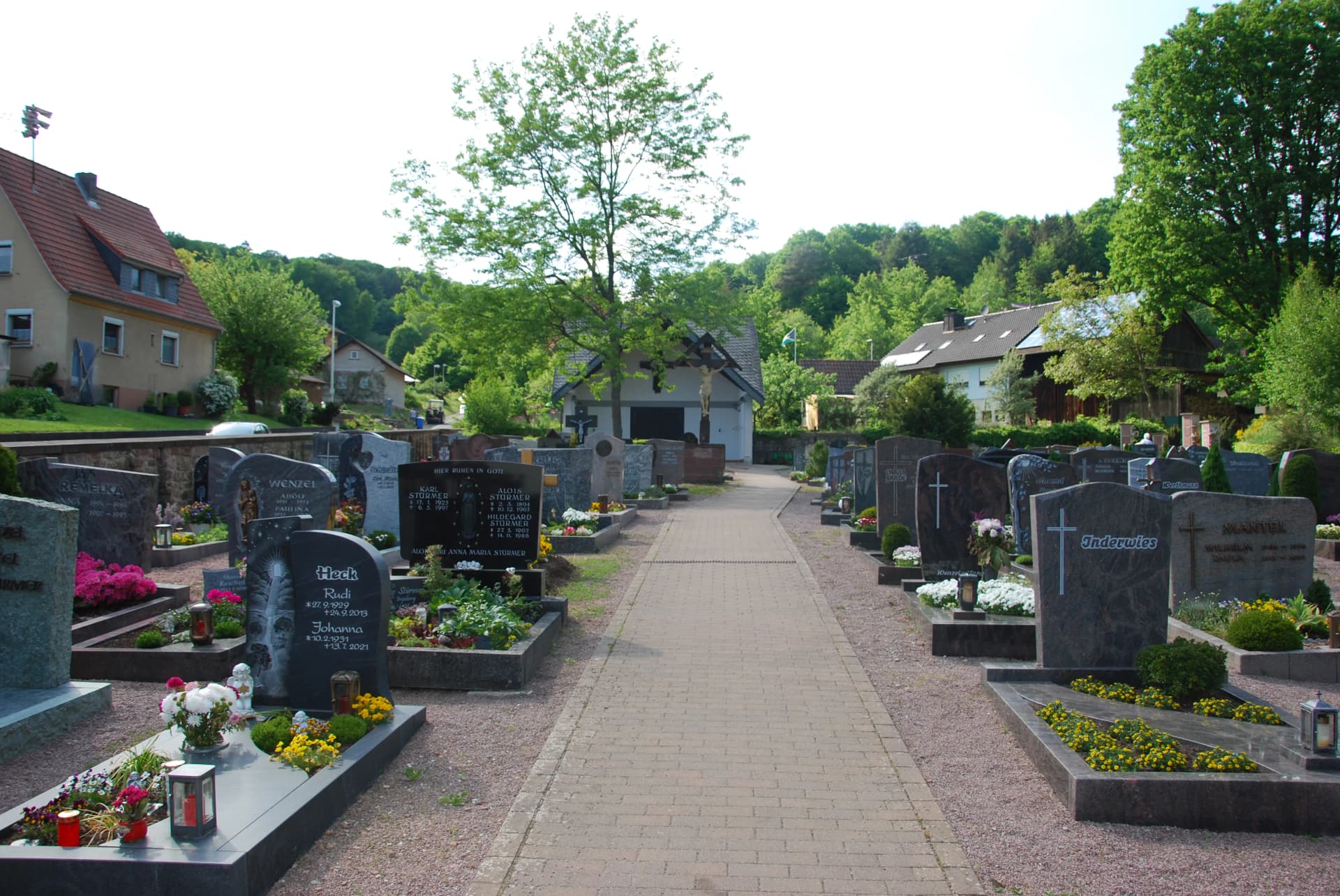 Friedhof mit gepflegten Grabsteinen und Blumenschmuck entlang eines gepflasterten Wegs, im Hintergrund Wohnhäuser und Wald