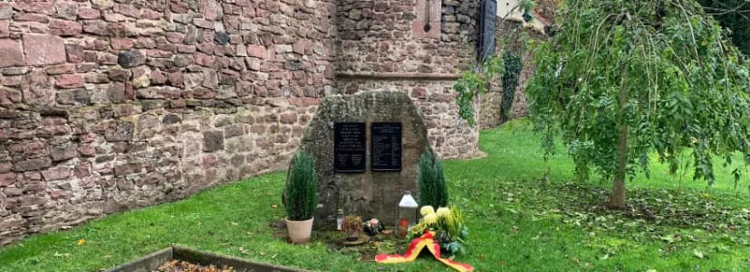 Gedenkstein mit zwei Gedenktafeln vor einer alten Steinmauer, umgeben von Grün, Kerzen und Blumen