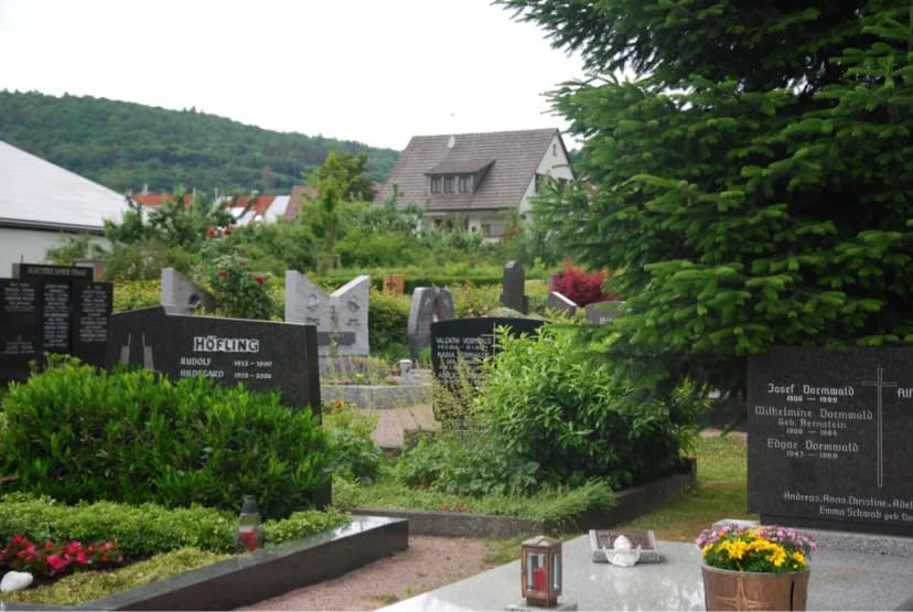 Friedhof mit Grabsteinen und bepflanzten Grabbeeten, im Hintergrund Wohnhäuser und ein bewaldeter Hügel.