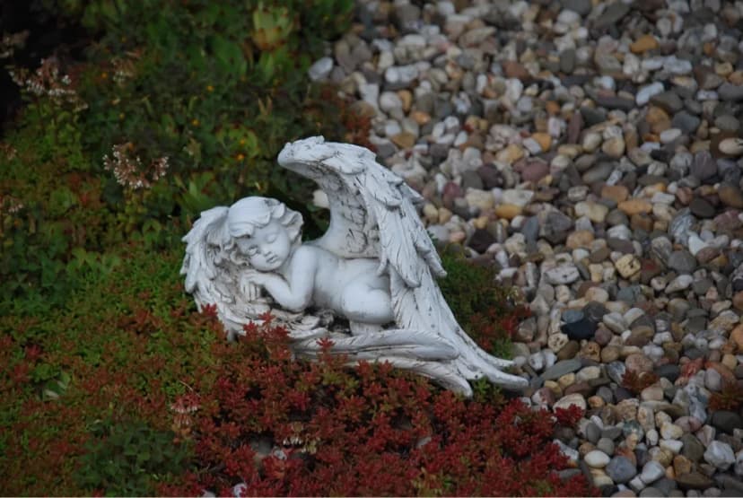 Steinfigur eines liegenden Engels zwischen Bodendecker und Kieselsteinen im Garten