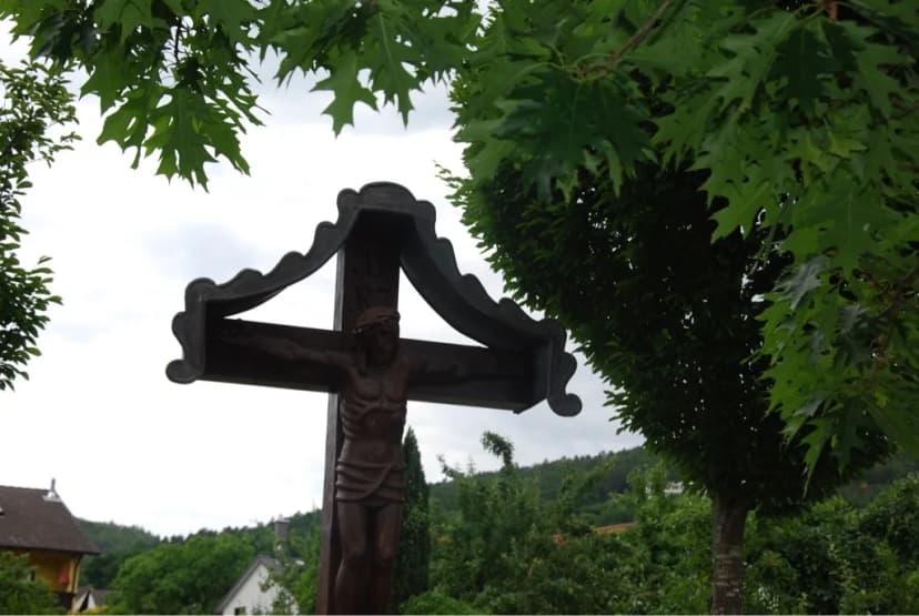 Hölzernes Wegkreuz mit geschnitzter Christusfigur und Schutzdach, umgeben von Bäumen