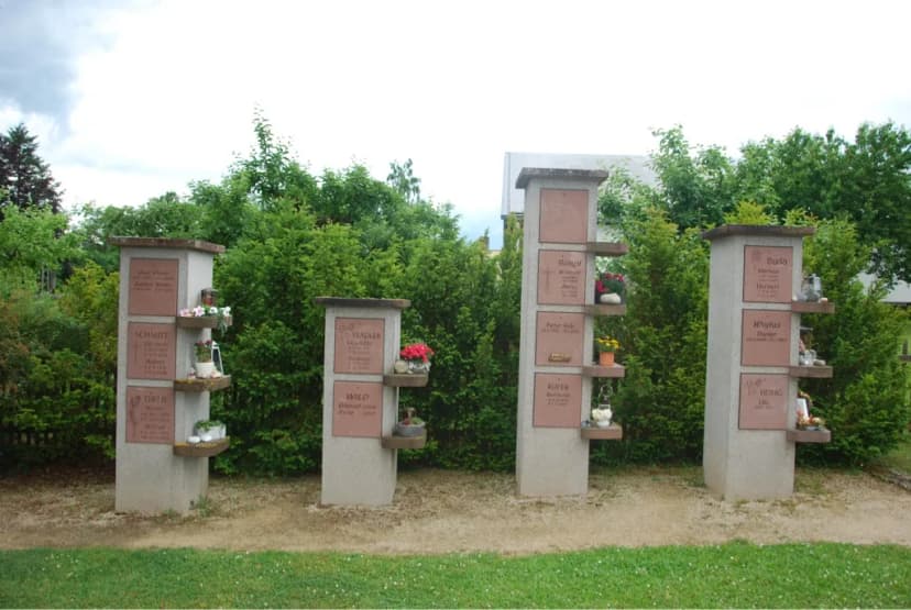 Vier steinerne Grabstelen mit Namenstafeln und kleinen Ablagen für Blumen und Grabkerzen auf einem Friedhof vor einer Hecke.