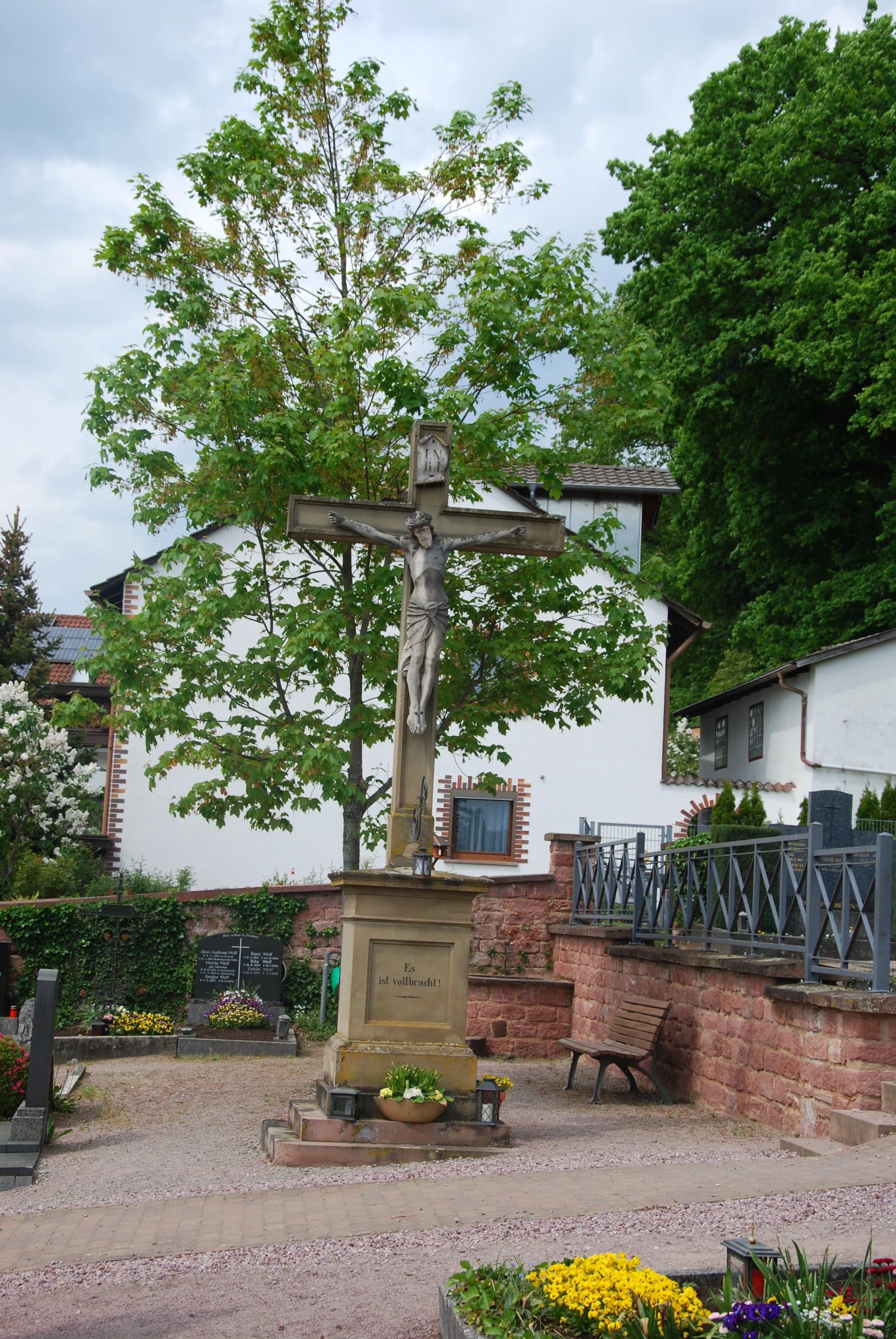 Steinernes Kreuz mit Christusfigur auf einem Friedhof, umgeben von Bäumen und Blumenbeeten.