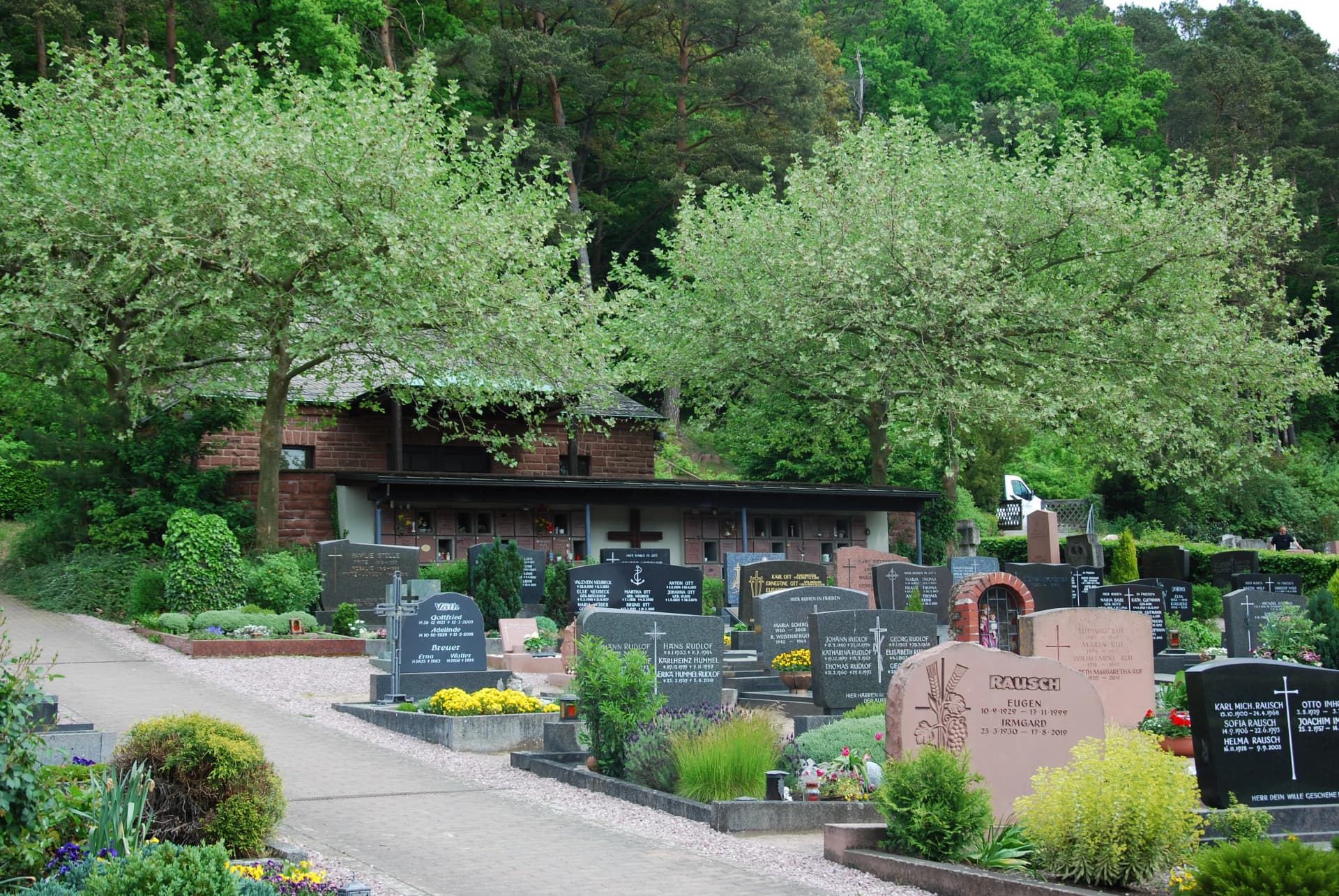 Friedhof mit Grabsteinen, bepflanzten Gräbern und einer kleinen Kapelle unter Bäumen