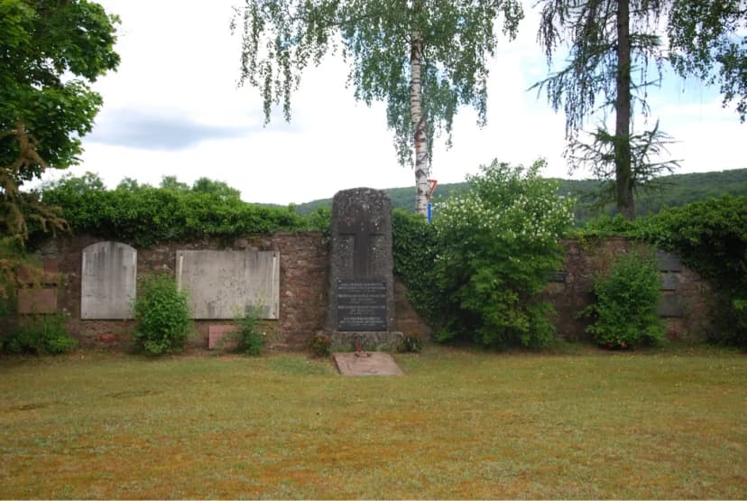 Gedenkstein mit Kreuz und Inschrifttafeln vor einer efeubewachsenen Friedhofsmauer mit weiteren Gedenktafeln