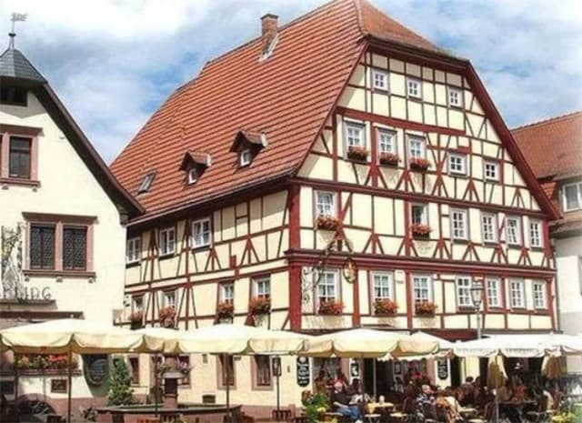 Fachwerkhaus mit rotem Ziegeldach und Blumenschmuck, davor Sonnenschirme eines Straßencafés