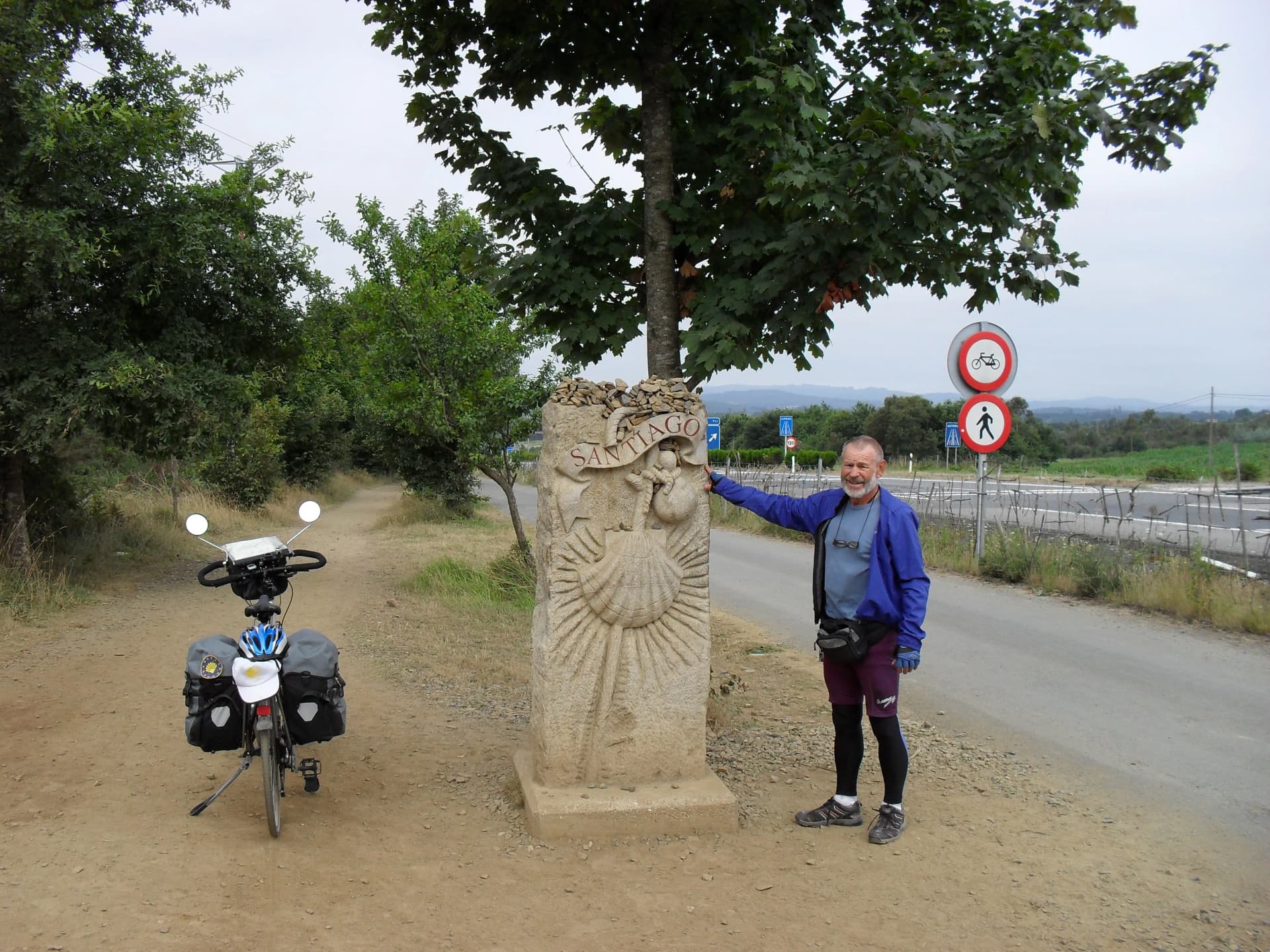 Mann steht lächelnd neben einem Steinmal mit Muschelsymbol und Aufschrift „Santiago“, daneben ein bepacktes Fahrrad, im Hintergrund Bäume und eine Landstraße.