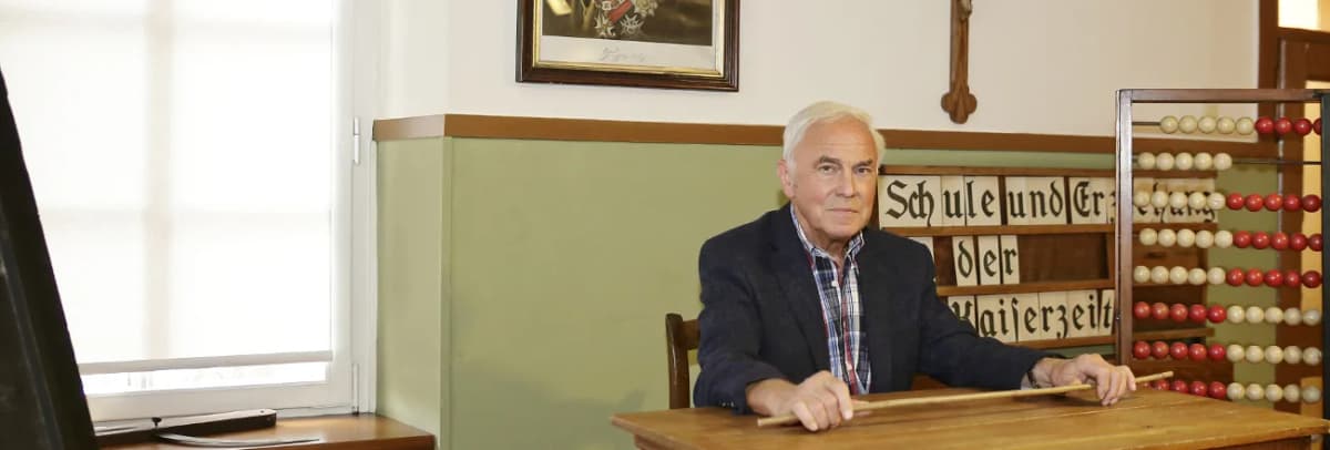 Mann sitzt an einem alten Schulpult mit Zeigestock, im Hintergrund ein historisches Rechenbrett und eine Schrifttafel zu Schule und Erziehung