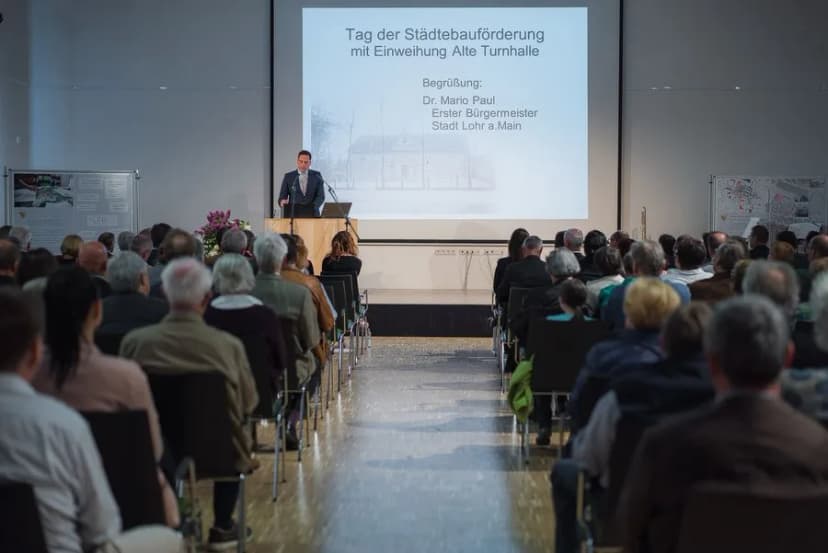 Bürgermeister spricht am Rednerpult vor Publikum, im Hintergrund Leinwand mit Titel Tag der Städtebauförderung mit Einweihung Alte Turnhalle