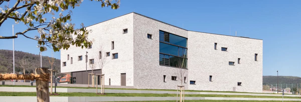 Moderner Gebäudekomplex mit heller Klinkerfassade und unregelmäßig angeordneten Fenstern, umgeben von Rasenflächen und jungen Bäumen.