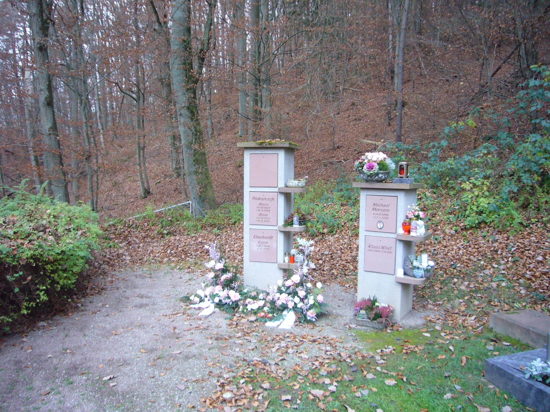 Zwei steinerne Urnenstelen mit Namensplatten, Blumenschmuck und Grablichtern vor einem herbstlichen Waldhang