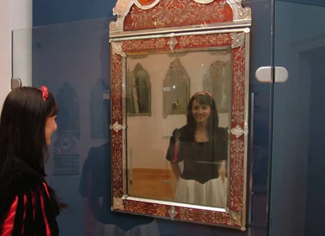 Junge Frau in historischem Kostüm blickt in einen verzierten Spiegel mit rotem Rahmen im Museum.