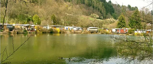 Teich mit Enten, umgeben von Wohnwagen und Hütten am Waldhang