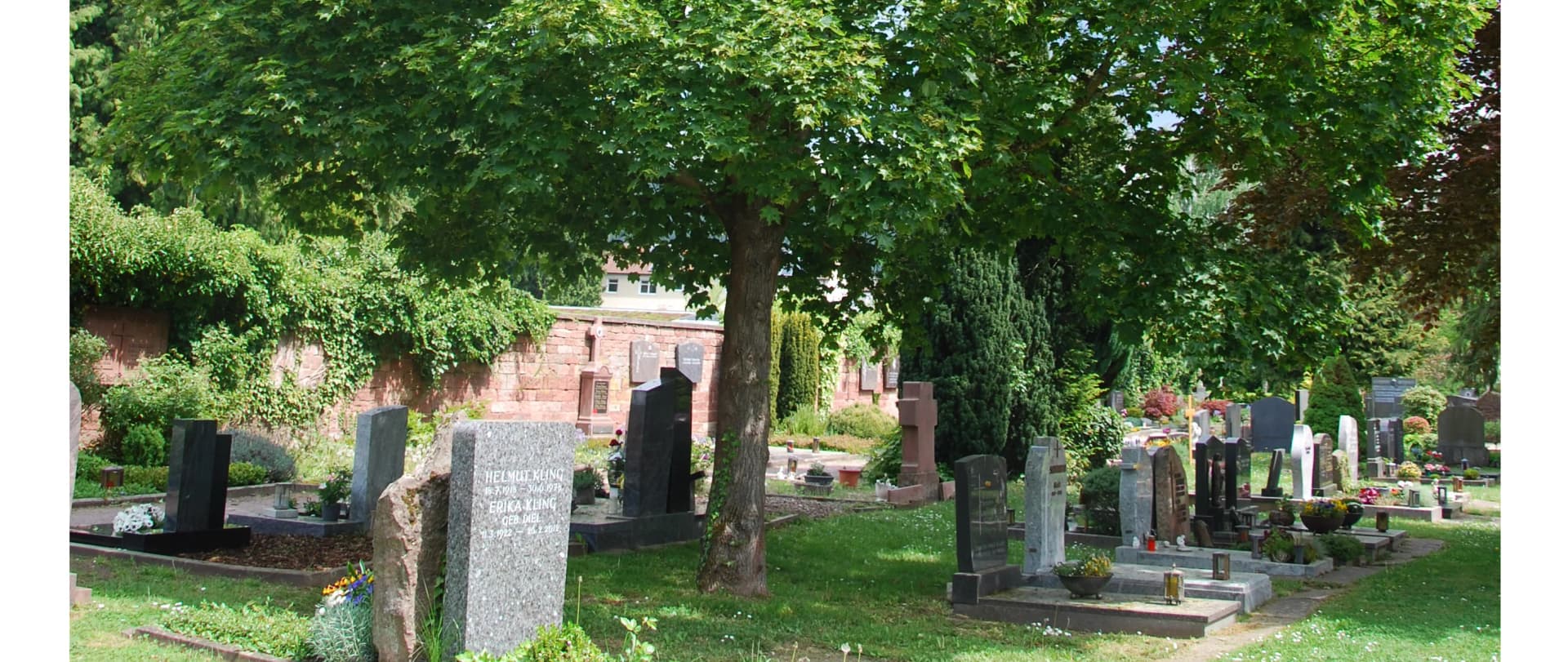 Friedhof mit Grabsteinen unter grünen Bäumen, umgeben von einer Backsteinmauer