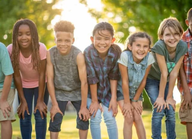 Gruppe lachender Kinder steht nebeneinander im Freien, nach vorne gebeugt mit Händen auf den Knien