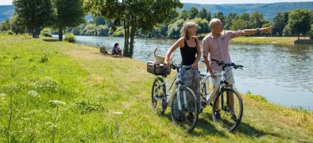 Zwei Radfahrende stehen mit ihren Fahrrädern am grünen Flussufer und schauen auf das Wasser