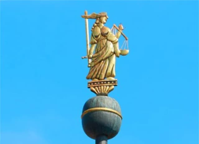 Goldene Justitia-Figur mit Waage und Schwert auf einer Turmspitze vor blauem Himmel