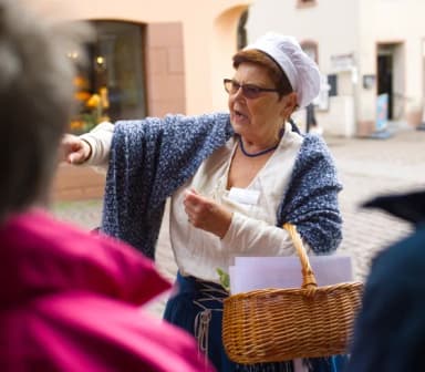 Eine als Wirtin kostümierte Stadtführerin erzählt über die Lohrer Stadtgeschicht. Verschwommen sind ein paar Zuhörer der Stadtführung zu sehen.