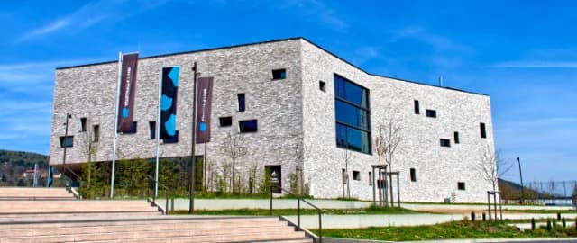 Moderne Stadthalle mit heller Steinfassade und unregelmäßig angeordneten Fenstern, davor Fahnen und eine Treppe