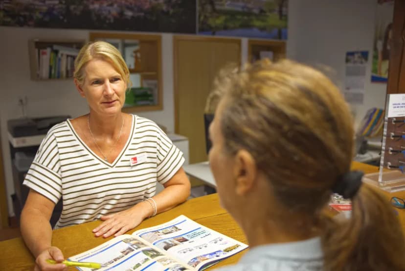 Eine Frau mit braunen Haaren wird in der Touristinformation von einer Mitarbeiterin beraten
