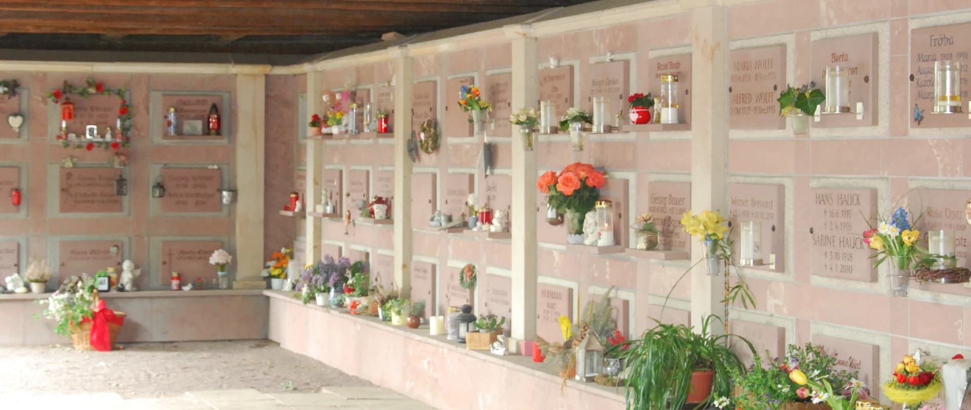Überdachte Urnenwand mit zahlreichen Grabnischen, geschmückt mit Blumen, Kerzen und Namensschildern