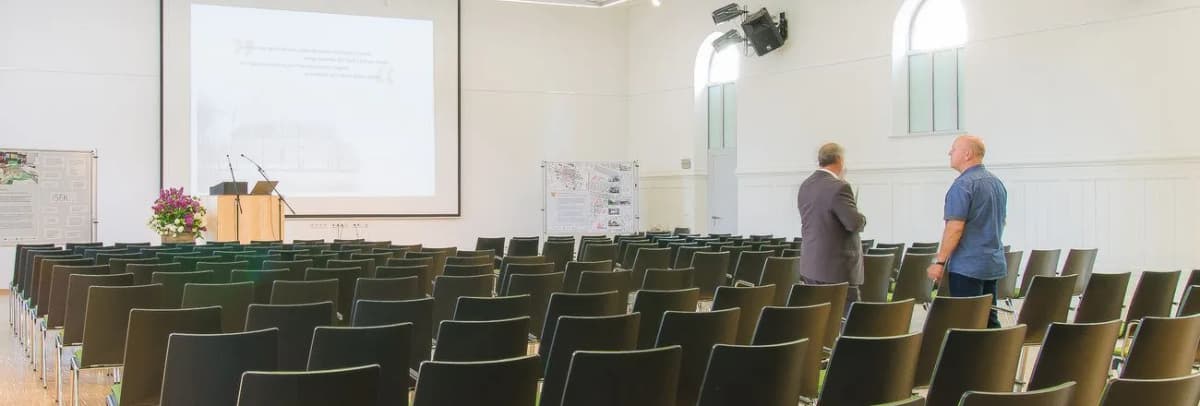 Leerer Veranstaltungssaal mit Stuhlreihen, Rednerpult und Projektionsleinwand, zwei Männer stehen im Hintergrund