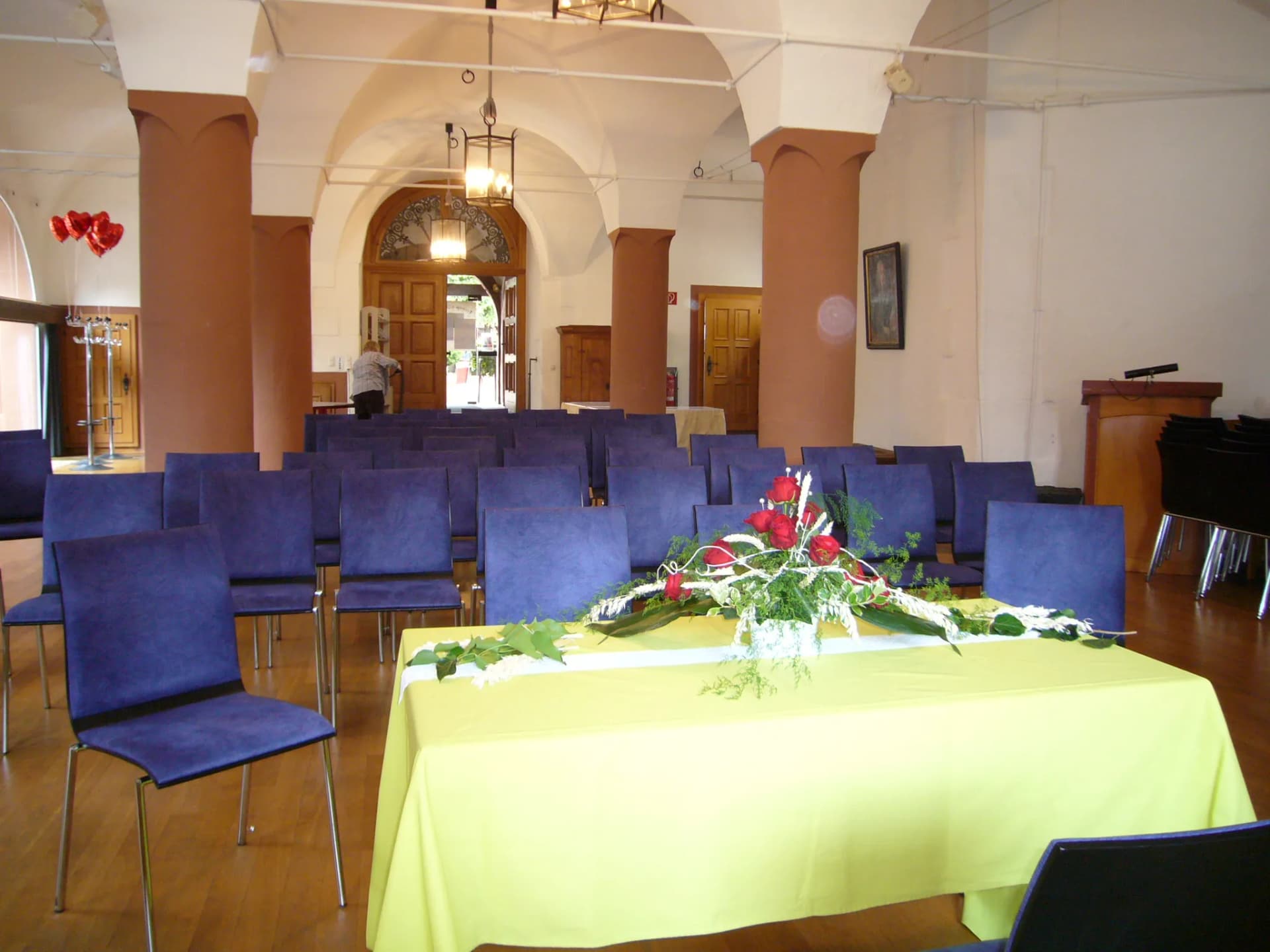 Gewölbesaal mit Reihen blauer Stühle und einem Tisch mit gelber Decke und Blumengesteck aus roten Rosen im Trauraum des Alten Rathauses.