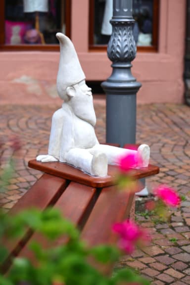 Blick auf eine der Zwergen-Figuren, die auf einer Parkbank in der Lohrer Innenstadt sitzt. Im Vordergrund sind Blumen zu sehen.