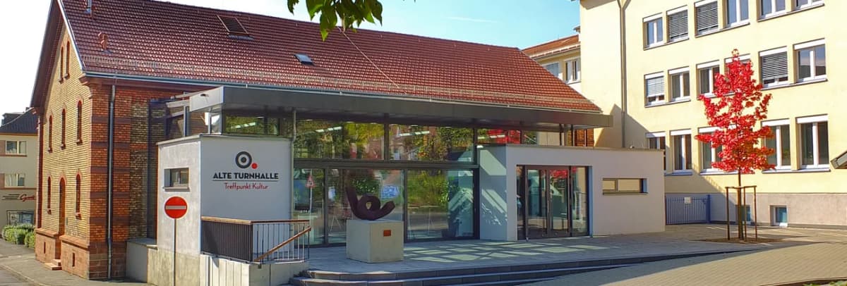 Gebäude der Alten Turnhalle mit rotem Ziegeldach, gläsernem Vorbau und Schriftzug 'Alte Turnhalle – Treffpunkt Kultur'