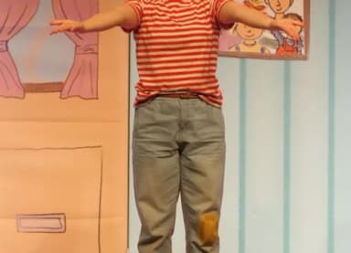 Person im rot-weiß gestreiften Shirt und Jeans mit ausgebreiteten Armen auf einer Theaterbühne mit gemalter Kulisse