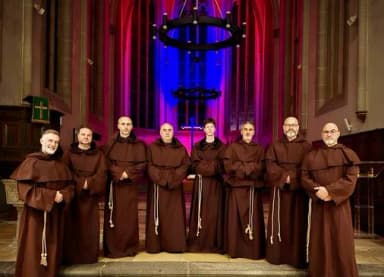 Acht Mönche in braunen Kutten stehen nebeneinander in einer Kirche mit farbig beleuchtetem Altarraum.