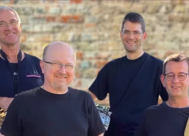 Vier lächelnde Männer in dunklen T-Shirts stehen vor einer Mauer