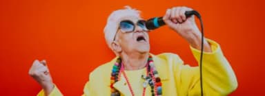 Ältere Frau in gelber Jacke singt mit Mikrofon vor orangem Hintergrund