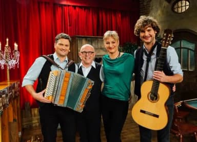Vier Personen stehen vor einem roten Vorhang, ein Mann hält ein Akkordeon, ein anderer eine Gitarre