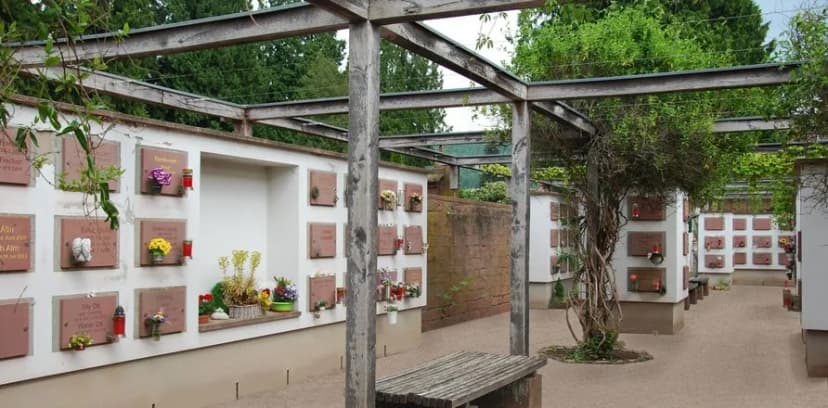 Urnenwand auf einem Friedhof mit Gedenktafeln, Blumen und Grablichtern unter einer Pergola