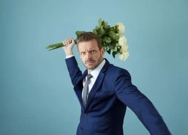 Mann im blauen Anzug hält sich einen Blumenstrauß mit weißen Rosen an den Kopf und blickt grimmig