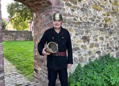 Mann in historischer Feuerwehruniform mit Messinghelm hält einen alten Löscheimer vor einer Steinmauer.