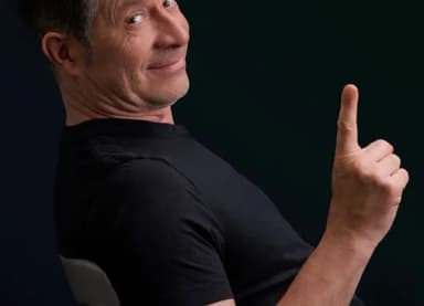 Mann in schwarzem T-Shirt blickt lächelnd über die Schulter und hebt den Zeigefinger