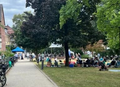 Menschen sitzen und stehen auf einer Parkwiese unter Bäumen bei einem Freiluftveranstaltung mit Pavillonzelten im Hintergrund.