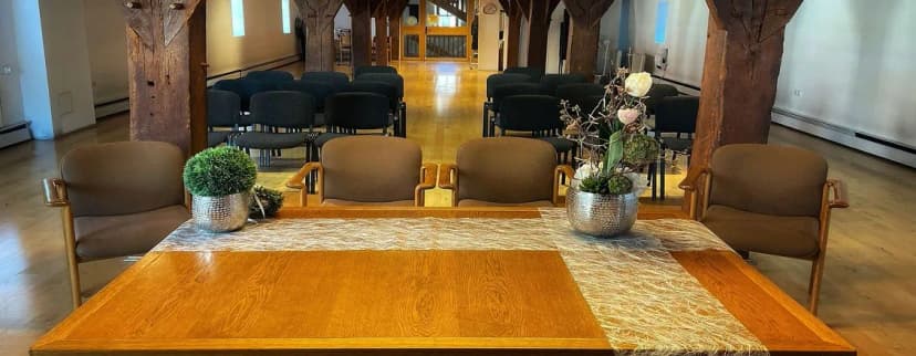 Innenraum der Kellereischeune mit Holzbalken, Stuhlreihen und einem Tisch mit Blumendekoration im Vordergrund
