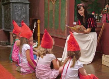 Kostümierte Frau liest Kindern mit roten Zipfelmützen aus einem Bilderbuch vor