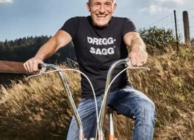Mann mit T-Shirt „Dregg-Sagg“ sitzt lachend auf einem umgebauten Fahrrad auf einer Wiese