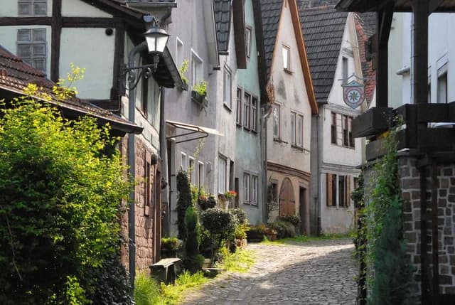 Enge Gasse mit gepflastertem Boden zwischen alten Fachwerk- und Giebelhäusern, an einer Hauswand hängt eine schmiedeeiserne Laterne