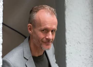 Porträt eines Mannes mit kurzem grauem Haar und Bart in grauem Sakko, der aus einem Torbogen blickt