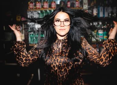 Frau mit schwarzen langen Haaren, Brille und Leopardenmuster-Kleid vor einer Bar