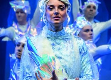 Bühnendarsteller in glitzernden, eisblauen Kostümen mit weißen Perücken, im Vordergrund eine Frau mit kristallartigem Requisit