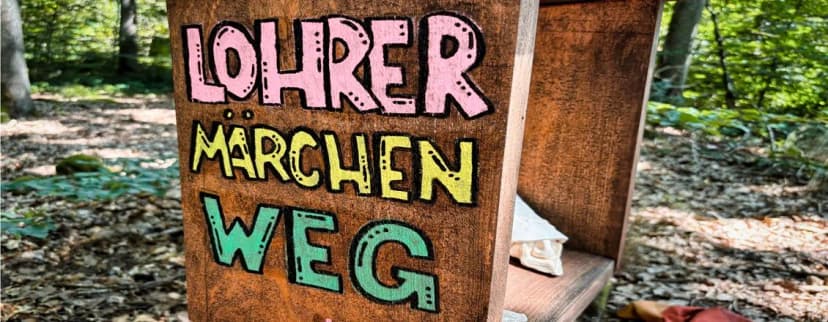 Holzschild mit bunter Aufschrift „Lohrer Märchenweg“ im Wald
